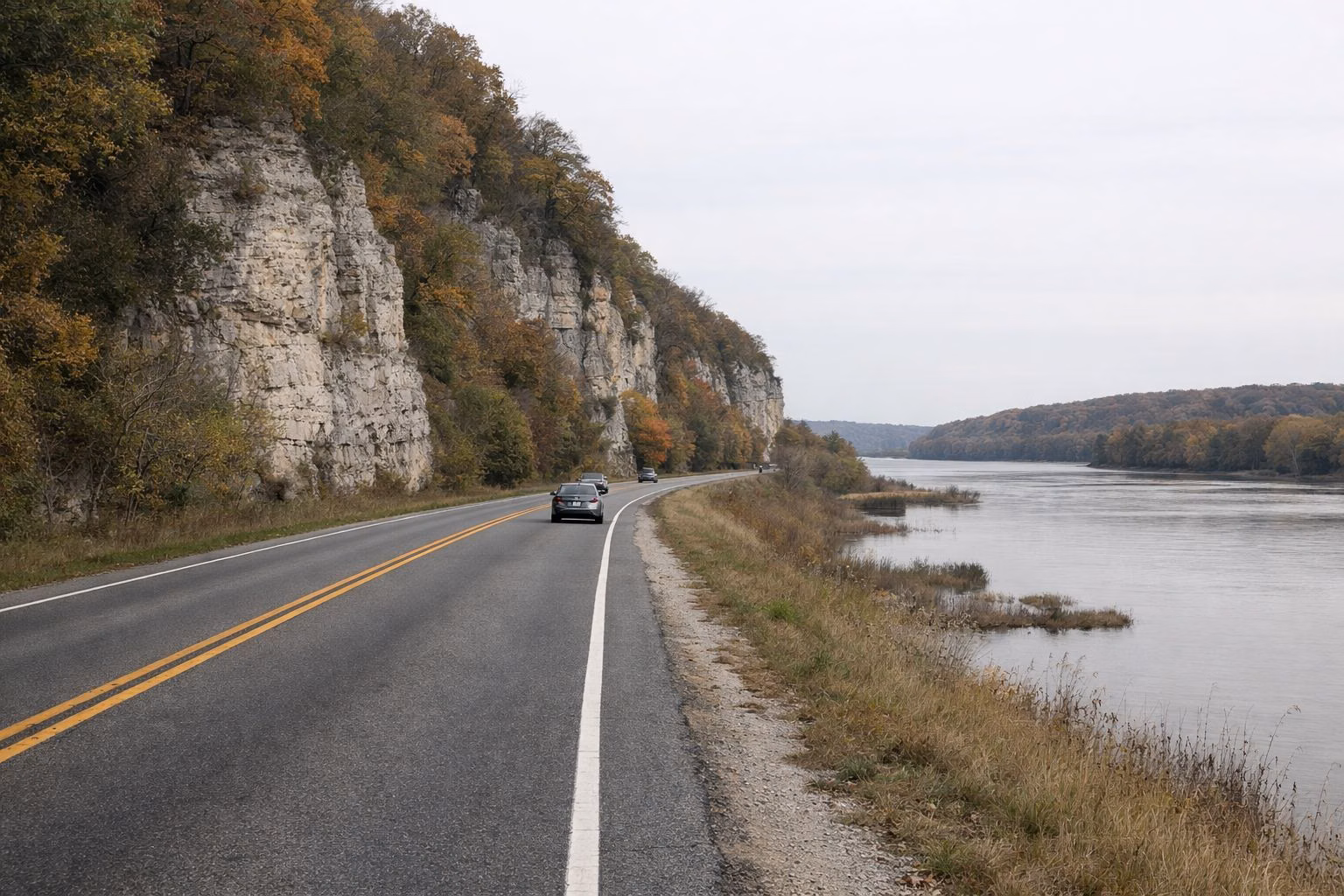 A two-lane highway running along the base of tall pale limestone bluffs with a wide river on the other side