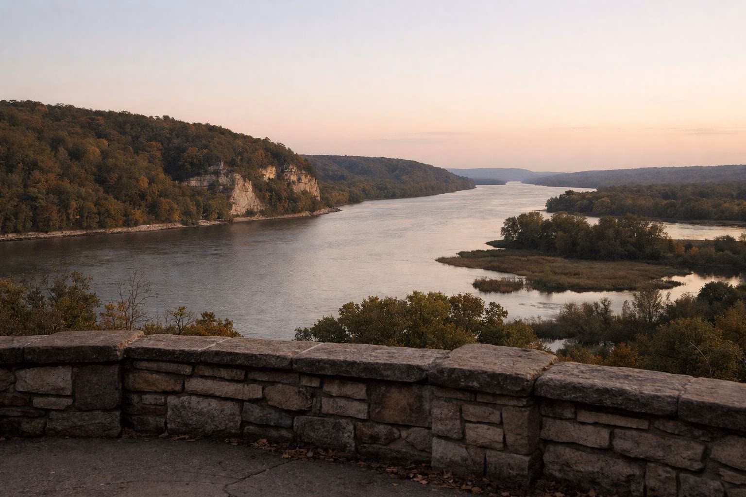 A low stone wall terrace at dusk overlooking a wide river and wooded hills, warm last light