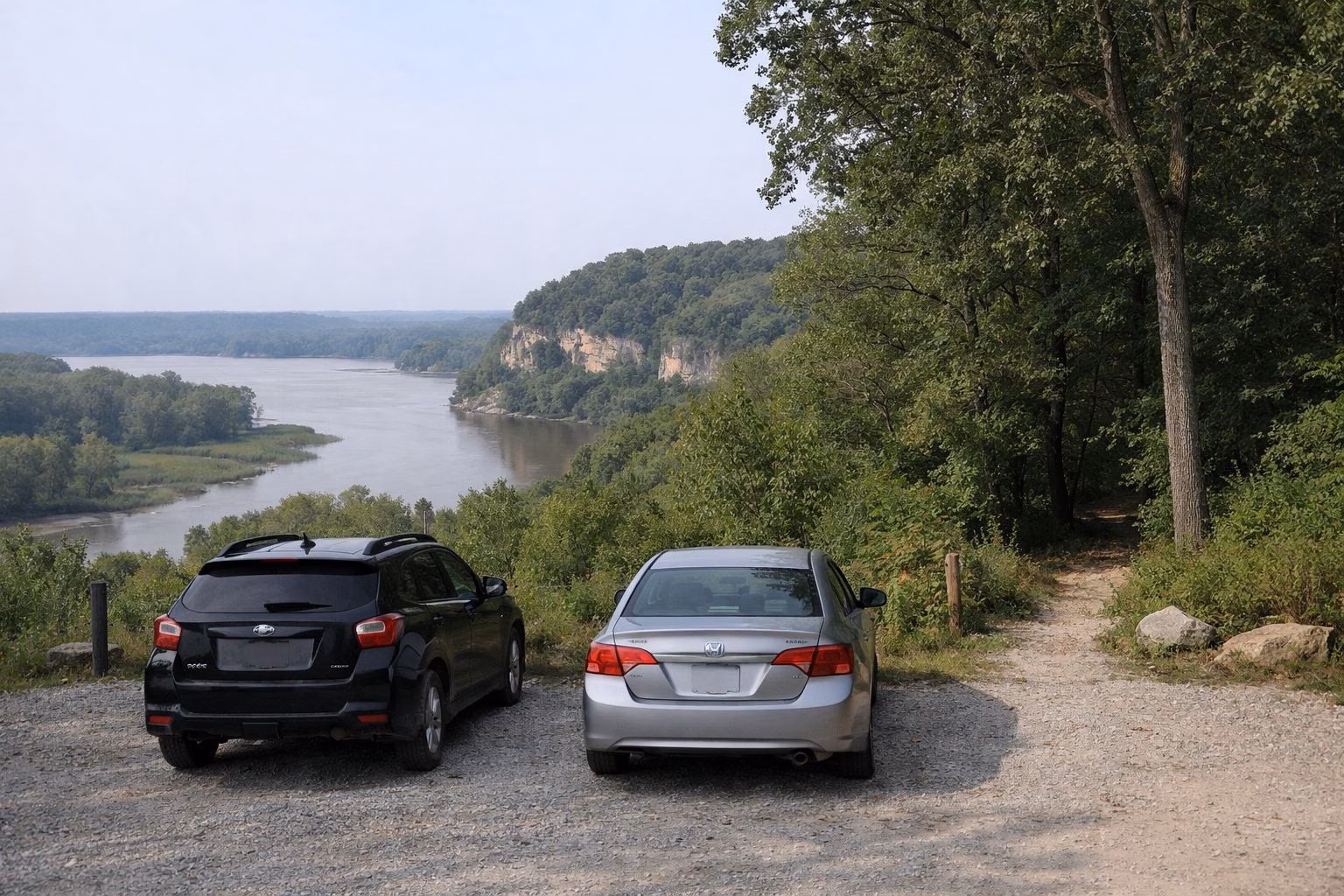 A small gravel parking area at the edge of an oak forest with a trail entering the woods, two parked cars seen from behind
